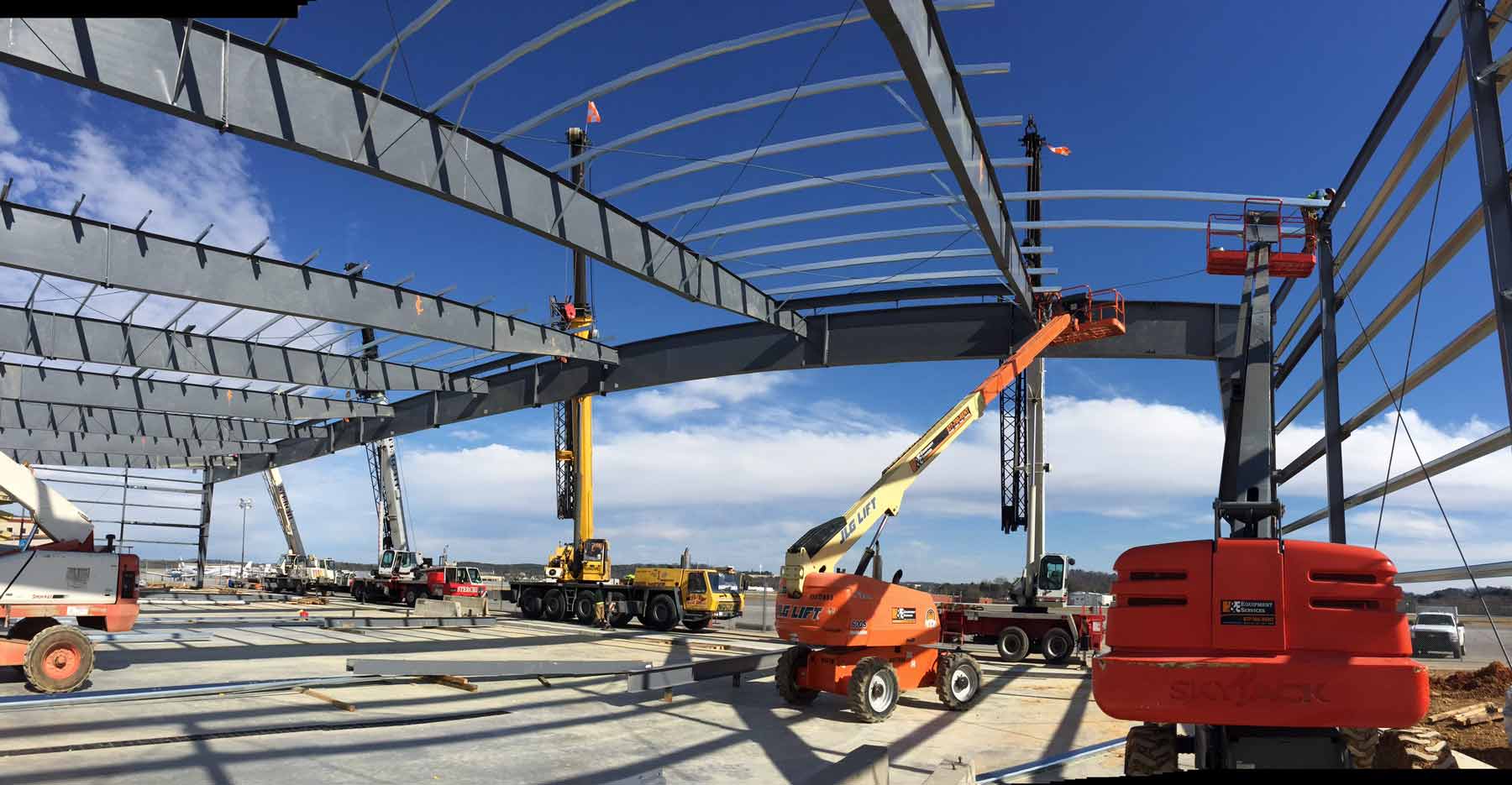 Steel framing going up on a structural hangar project with cranes and lifts setting large beams against a clear blue sky.