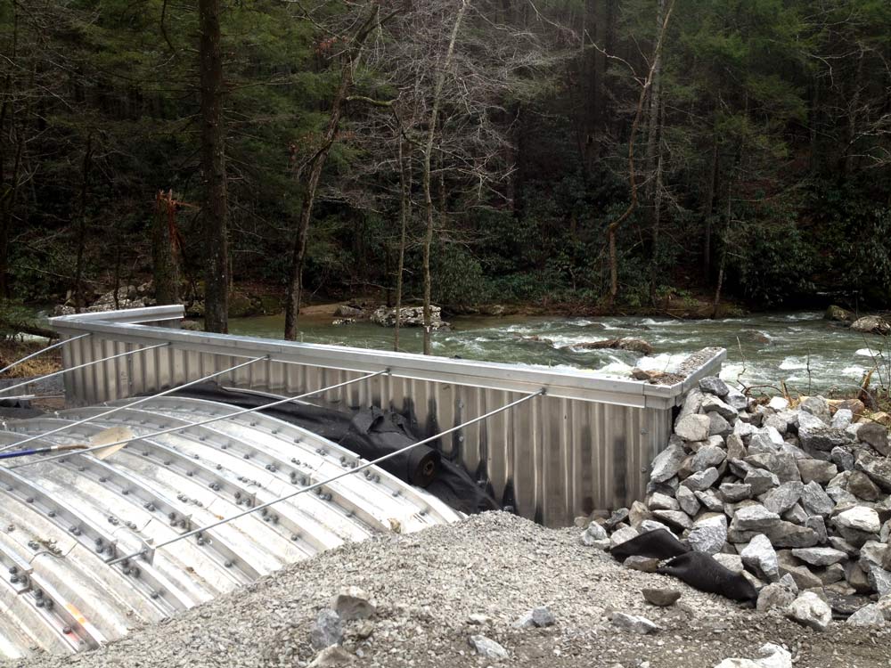 A metal culvert channels a small stream beneath a gravel road in the Cherokee National Forest in Polk County, Tennessee. Rock riprap lines the banks, and water flows steadily through the new crossing within a wooded setting.