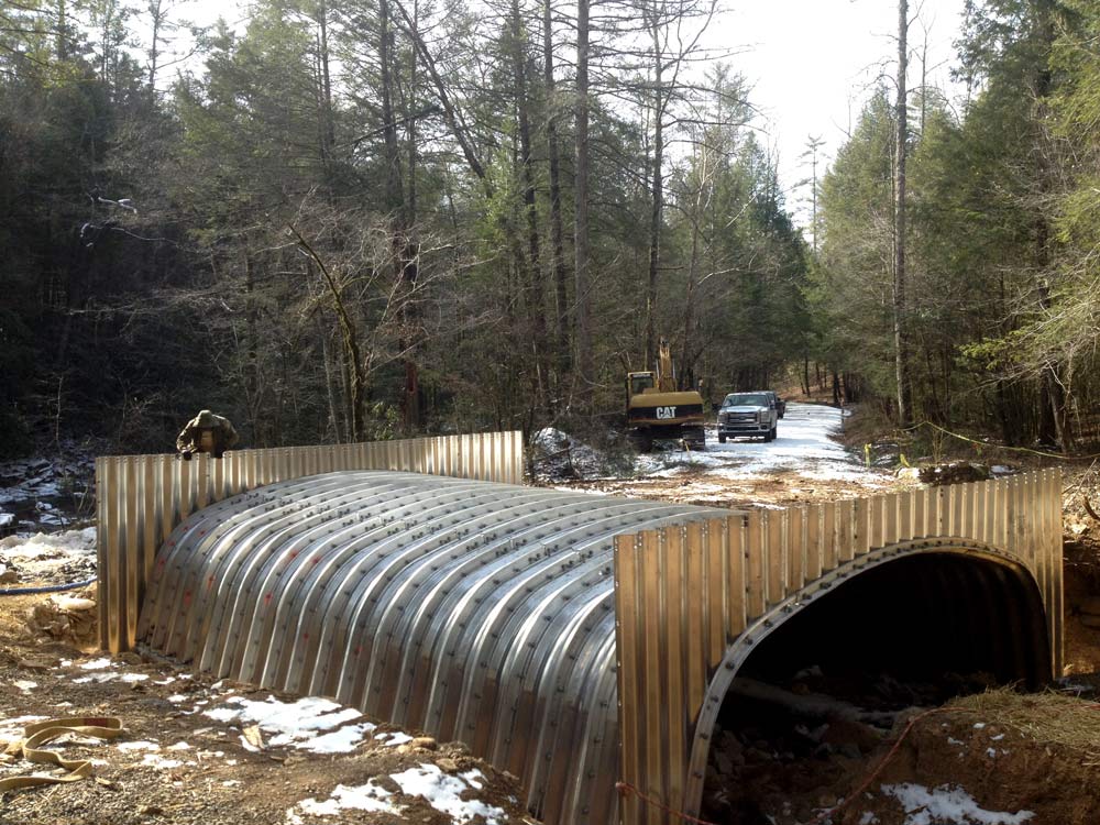 A metal culvert channels a small stream beneath a gravel road in the Cherokee National Forest in Polk County, Tennessee. Rock riprap lines the banks, and water flows steadily through the new crossing within a wooded setting.
